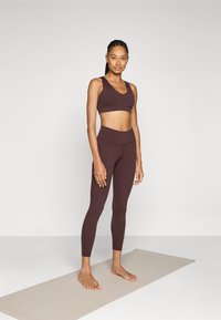 Rjavi športni crop top in visokorzni legingi iz raztegljivega materiala, ki se ponašajo z gladko teksturo in prilegajočim dizajnom.