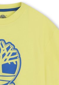 T-shirt di cotone gialla con una grande grafica di un albero blu sul davanti. Collo a giro con colletto interno blu a contrasto.
