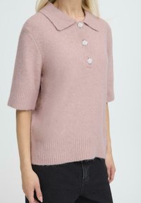 Pull en tricot rose pâle à manches courtes avec un col et trois boutons décoratifs. Présente un ourlet côtelé et une texture douce.