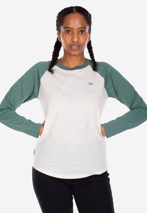 Young woman with braided hair wearing a white and green long-sleeve raglan shirt and black pants, standing with hands on hips.