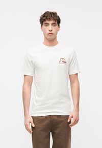 Νεαρός άνδρας με καστανά σγουρά μαλλιά που φοράει λευκό T-shirt με ένα μικρό κόκκινο λογότυπο κύματος και βουνού στην αριστερή πλευρά του στήθους, συνδυασμένο με καφέ παντελόνι.