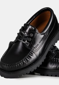 Scarpe in pelle nera con punta arrotondata, cuciture visibili e suola testurizzata. Presentano lacci e un marchio in rilievo sul lato.