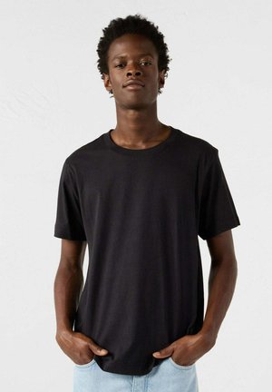 Zwarte katoenen t-shirt met ronde hals, korte mouwen en een losse pasvorm, gecombineerd met lichtblauwe jeans voor een neutrale achtergrond.