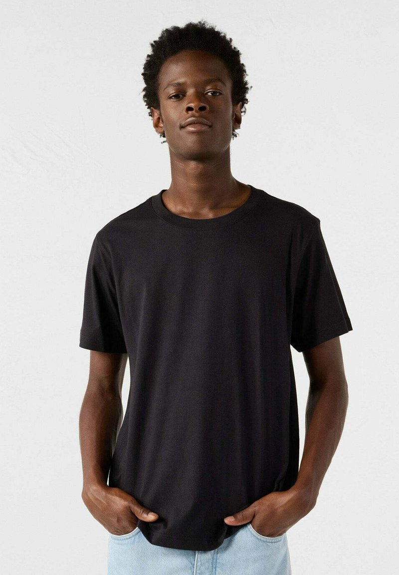 T-shirt nera in cotone con colletto rotondo, maniche corte e un fit largo, abbinata a jeans azzurri chiari su uno sfondo neutro.