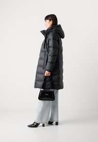 Abrigo acolchado negro con capucha, cierre de cremallera y cordones elásticos. Combinado con jeans de pierna ancha azul claro y un bolso negro.