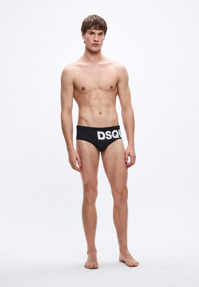 Μαύρα μαγιό σλιπ με υφασμάτινο μοτίβο και φαρδιά ζώνη με έντονη λευκή επιγραφή "DSQUARED2". Το μοντέλο στέκεται ξυπόλυτο μπροστά σε απλό φόντο.