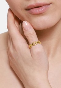 Bague en chaîne en or portée à un doigt, avec des maillons lisses et arrondis. Peau visible avec du vernis à ongles naturel sur les doigts.