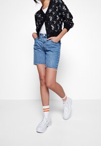 Nainen, jolla on päällä musta kuvioitu neuletakki, valkoinen paita, siniset farkkushortsit, valkoiset sukat oransseilla raidoilla ja valkoiset korkeavartiset lenkkarit, kädet taskuissa.