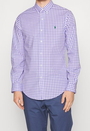 Man draagt een paars-wit geruit shirt met lange mouwen en een groen logo, gecombineerd met donkerblauwe broek, staand tegen een effen achtergrond.