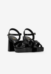 Sandalias de plataforma de charol negro con tacón cuadrado, que presentan múltiples correas entrelazadas y una correa ajustable en el tobillo.