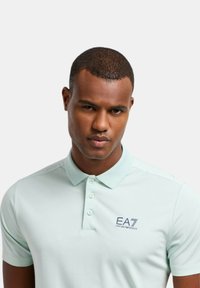 Polo de color verde menta claro hecho de una tela suave, con una tapeta de tres botones y el logotipo azul oscuro de EA7 en el lado izquierdo.