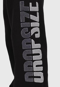 Schwarze Jogginghosen mit strukturiertem grauen "DROPSIZE" Schriftzug entlang des Beins, wobei unterschiedliche Stoffhöhen einen 3D-Effekt erzeugen.