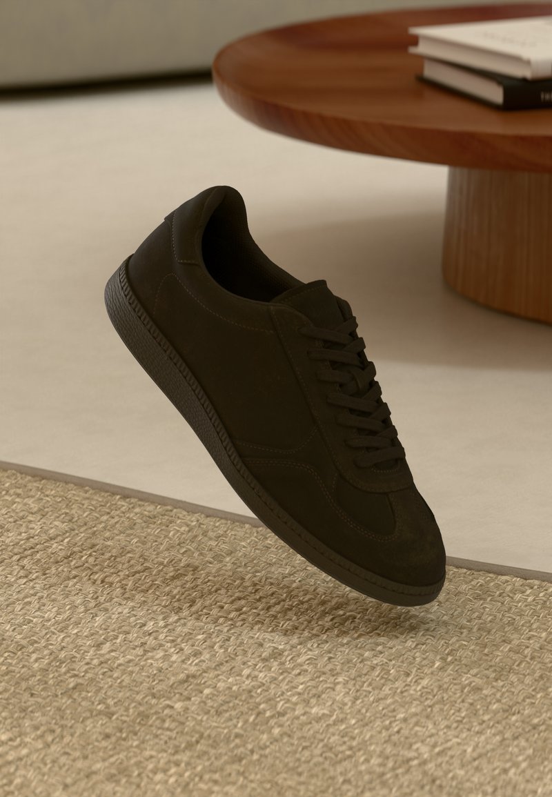 Črne semiš superge z zaobljenim nosom, z vezalkami, teksturiranim gumijastim podplatom in kontrastnim šivom, razstavljene nad prejo.