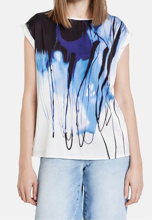 Woman wearing a loose white sleeveless top with blue and black abstract ink drip patterns, paired with light blue denim jeans.