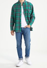 Camicia a quadri verde con bottoni sopra una canotta bianca, abbinata a jeans blu e sneakers bianche. La camicia presenta un colletto e una tasca sul petto.