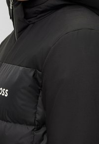 Svart puffjacka med en slät övre sektion och en texturerad nedre sektion, med en framdragkedja och logodetaljer i vitt.