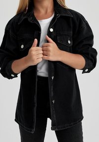 Chaqueta de mezclilla negra con cuello clásico, cierres de botón y bolsillos en el pecho. Mangas largas con puños enrollados, que presentan detalles en metal plateado.