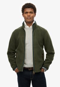 Giacca in pile verde con colletto alto, zip blu, tasche laterali, indossata sopra un maglione grigio, abbinata a jeans scuri e un orologio.