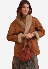 Vrouw in een bruine schapenvacht jas en een bloemenrok, met een roestkleurige suède trekkoordtas met studdetails.