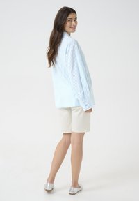 Camicia oversize a righe blu chiaro e bianche con maniche lunghe, abbinata a pantaloni corti beige e scarpe ballerine argentate. Tessuto di una consistenza morbida.