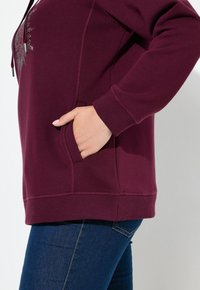 Felpa mauve con tasca frontale e polsini a coste. Presenta un design grafico testurizzato sul petto. Indossata con jeans in denim scuri.