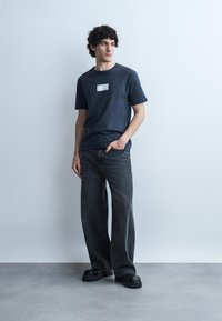 Jeune homme aux cheveux bouclés portant un T-shirt bleu marine, un jean noir ample et des chaussures noires épaisses, debout devant un mur gris clair uni.