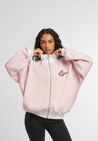 Felpa rosa con zip e dettagli bianchi, dotata di un logo frontale. Presenta una vestibilità oversized, tessuto morbido e colletto alto. Leggings neri indossati sotto.