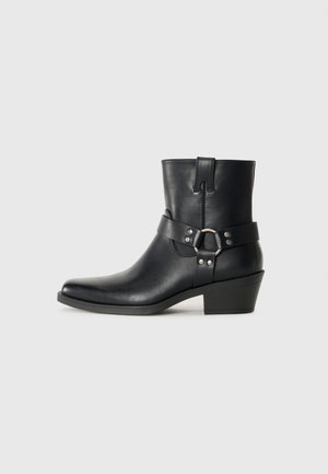 Bottines en cuir noir avec un bout pointu, une sangle décorative et une boucle, ainsi qu'un petit talon empilé. Texture lisse avec un minimum de détails.