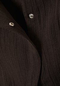 Brunet tekstil med en subtil bølget mønster. Har sølvknapper ved kragen, som tilfører funksjonelle detaljer til designet.