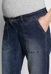 Jeans för gravida i denim med mörk tvätt, som har sidofickor, hög midja och kontrasterande guldstickningar. Mjuk tygtextur.