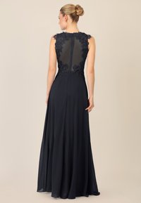 Abito da sera blu navy con dettagli in pizzo floreale, schiena trasparente e gonna fluente. Chiusura con zip sul retro, design senza maniche con bordi ondulati.