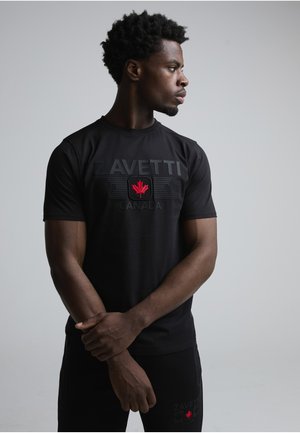 Mand i sort ZAVETTI Canada t-shirt og bukser, med armene krydset, ser mod højre mod en ensfarvet grå baggrund.