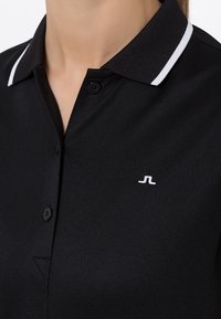 Černé polotričko s knoflíkovým zapínáním a bílým proužkovaným límečkem. Výrazné bílé logo na levé straně hrudi. Hladká textura.