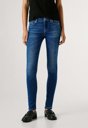 Persona vestida con vaqueros ajustados azul oscuro, mocasines negros y una camiseta negra, de pie frente a un fondo liso y claro.