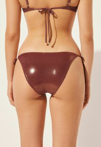 Bikiniunderdel i mörk burgundy, glänsande tyg med knyt-detaljer på sidan. Mjuk textur, måttlig täckning och figurnära design.