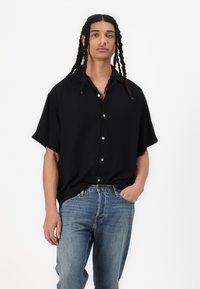 Schwarze kurzärmelige, locker geschnittene Hemdbluse, kombiniert mit blauen Jeans. Das Hemd hat einen Kragen und kontrastierende weiße Knöpfe.