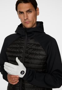 Man med en svart vadderad jacka med huva som justerar en vit golfhandske på sin vänstra hand mot en enkel bakgrund.