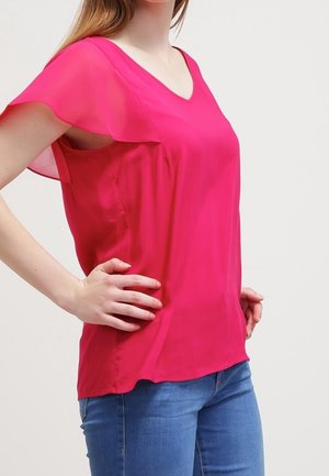 Roze V-halspolo met doorschijnende, losse mouwen, lichtgewicht stof en rechte snit. Gecombineerd met een blauwe spijkerbroek.