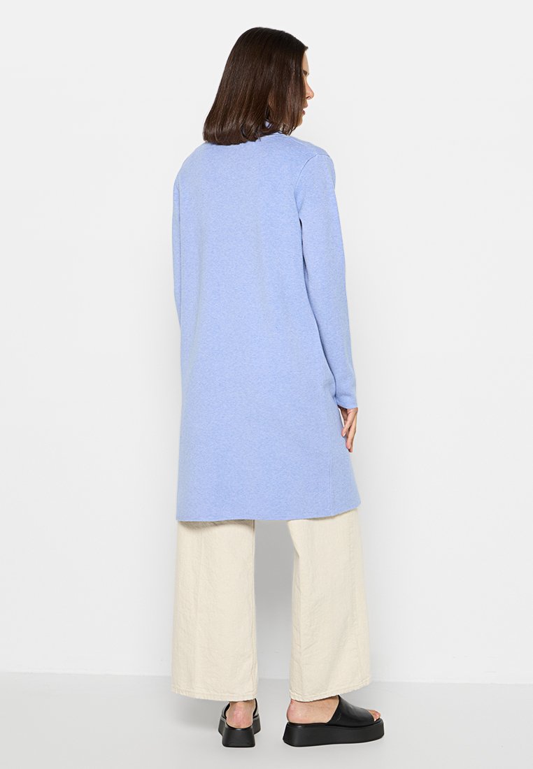 Langer, hellblauer Pullover mit geradem Schnitt und gerippten Bündchen, kombiniert mit beigefarbenen, weit geschnittenen Hosen und schwarzen Slippern.