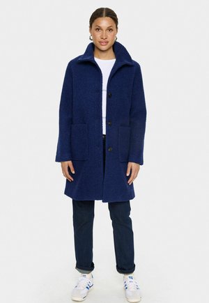 Marineblau strukturierter Mantel mit hohem Kragen, zwei Seitentaschen und Knopfverschluss, kombiniert mit dunklen Jeans und weißen Turnschuhen.