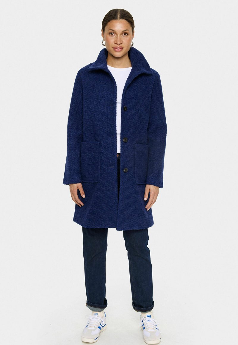 Marinblå strukturerad kappa med hög krage, två sidofickor och knappstängning, i kombination med mörka jeans och vita sneakers.