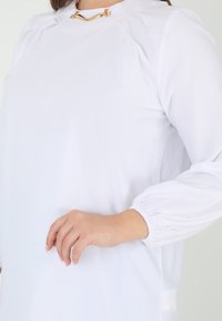 White blouse with a high neck, puffed long sleeves, and gathered detailing at shoulders. Features a gold-toned hardware accent at the neckline.