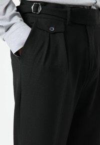 Zwarte broek met een gestructureerde stof, voorzien van plooien, een riem met een zilveren gesp en een knoopsluiting bij de zak.