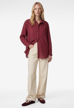 Lyshåret kvinde stående iført en løs, rødstribet skjorte med knapper, cremefarvede bukser med vide ben og bordeaux ballet flats med hvide snørebånd.