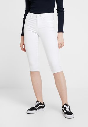 Person wearing white knee-length skinny pants, black long-sleeve top, and black-and-white sneakers standing on a white floor.