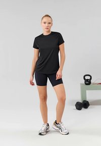 Crna sportska majica s kratkim rukavima u kombinaciji s crnim šortsevima. Model nosi srebrne i narančaste tenisice za trčanje, a u blizini su kettlebell i bučice.