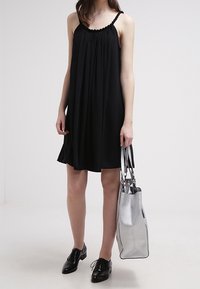 Black sleeveless dress with gathered neckline, paired with a silver tote bag and shiny black lace-up shoes. Smooth fabric, relaxed fit.