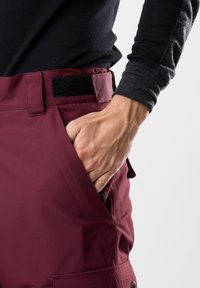 Burgundowe spodnie narciarskie z boczną kieszenią, wykonane z trwałego materiału o gładkiej teksturze. Zawierają czarne zapięcie na rzep i detale szycia.