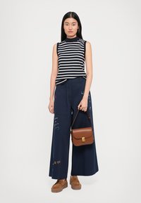 Pantaloni în stil larg de culoare bleumarin cu accente colorate scrise de mână, top fără mâneci cu dungi și geantă de umăr maro; încălțămintea are un design cu talpă groasă.