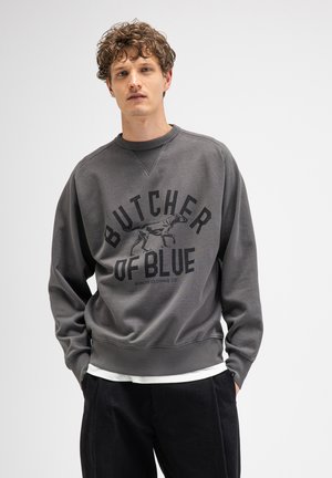 Young man with curly hair wearing a gray sweatshirt with "Butcher of Blue" and a dog graphic, standing against a plain light background.